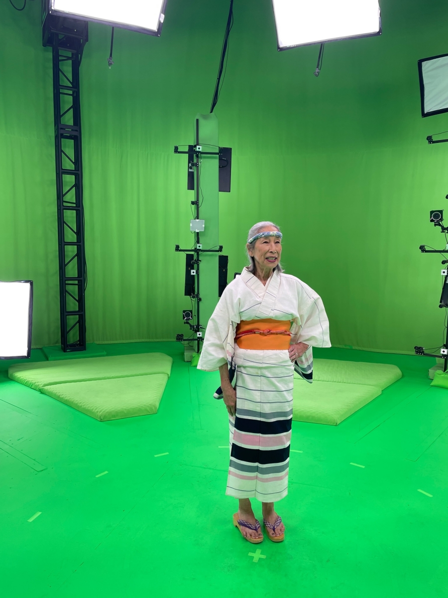 a woman dressed in Japanese kimono and headband stands in a green room filled with lights and cameras