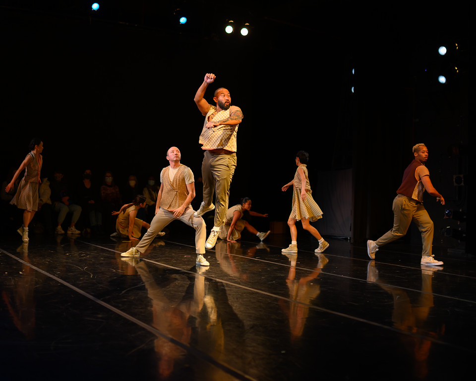 On a dark stage a small group of dancers are illuminated by light from above. One man is mid air, his arms raised, surrounded by the other dancers in mid-motion.