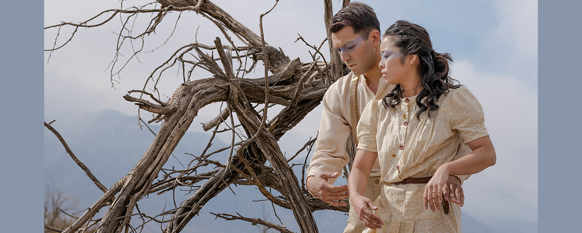 Two figures facing the camera but looking downward to the side, the man behind the woman with his arms around her waist. Twisted, gnarled, bare branches are next to them. Behind them is a range of mountains and smoky blue sky and clouds.