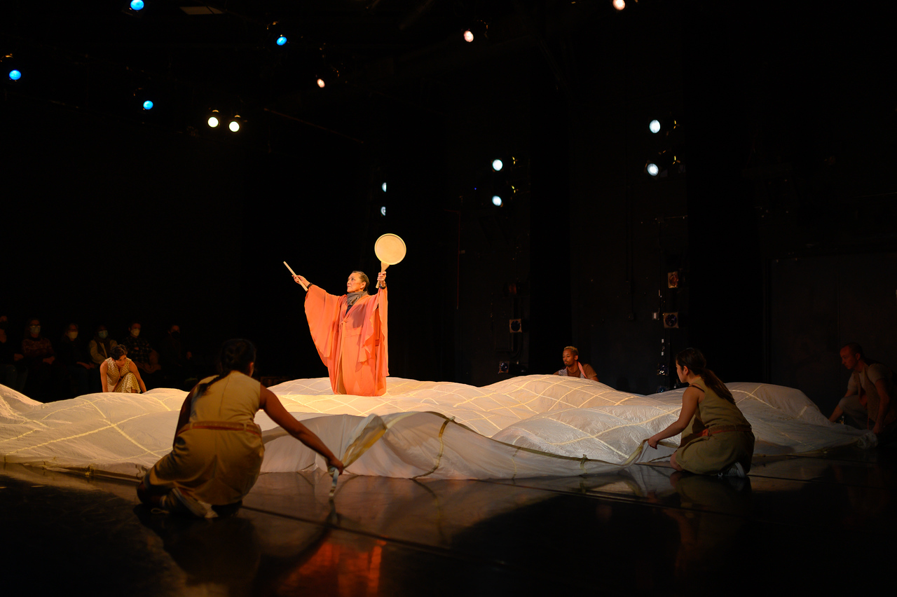 A woman in orange stands in the center of a large piece of white fabric, a fan in her upraised arm