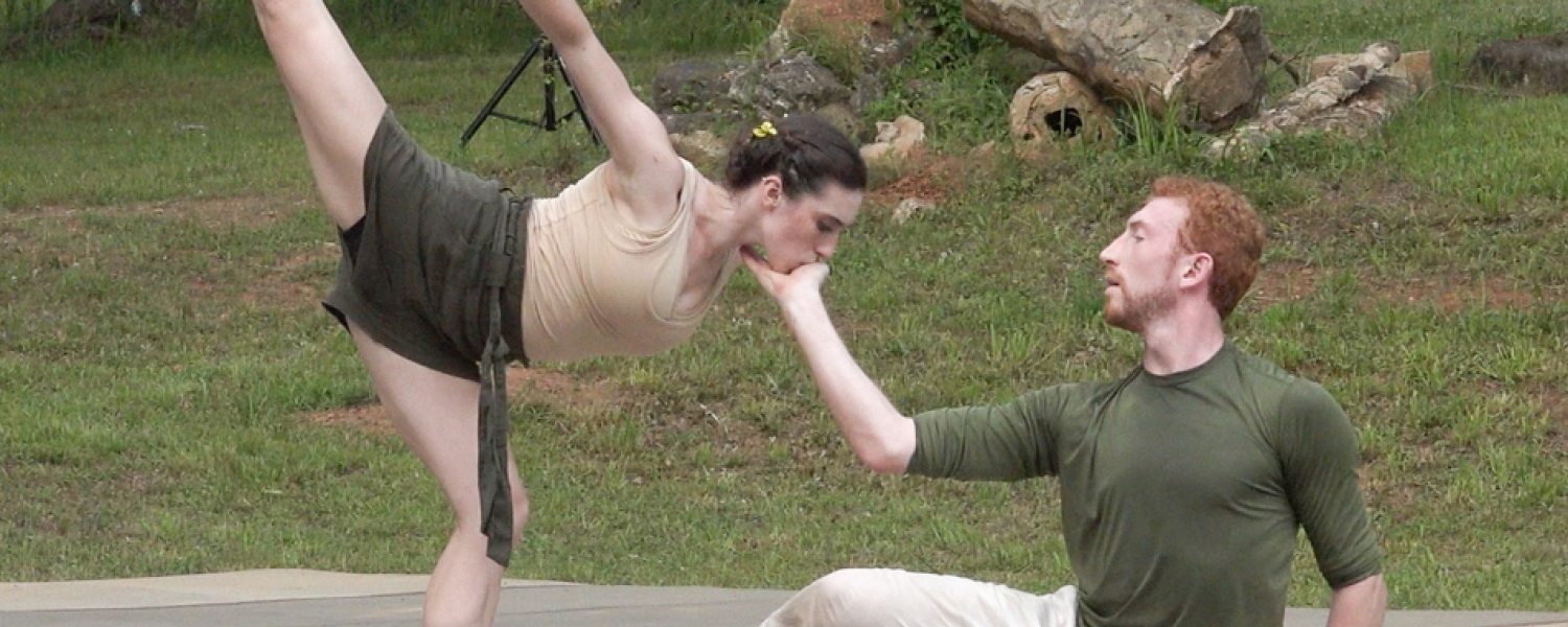 The setting is an outdoors, the woods and grass surrounding a low stage. A male dancer sits, one hand touching the ground, the other gently cupping the female dancer's face. She is posed in a deep arabesque towards the man, her face in his hand and her upper arm and leg extended skyward.