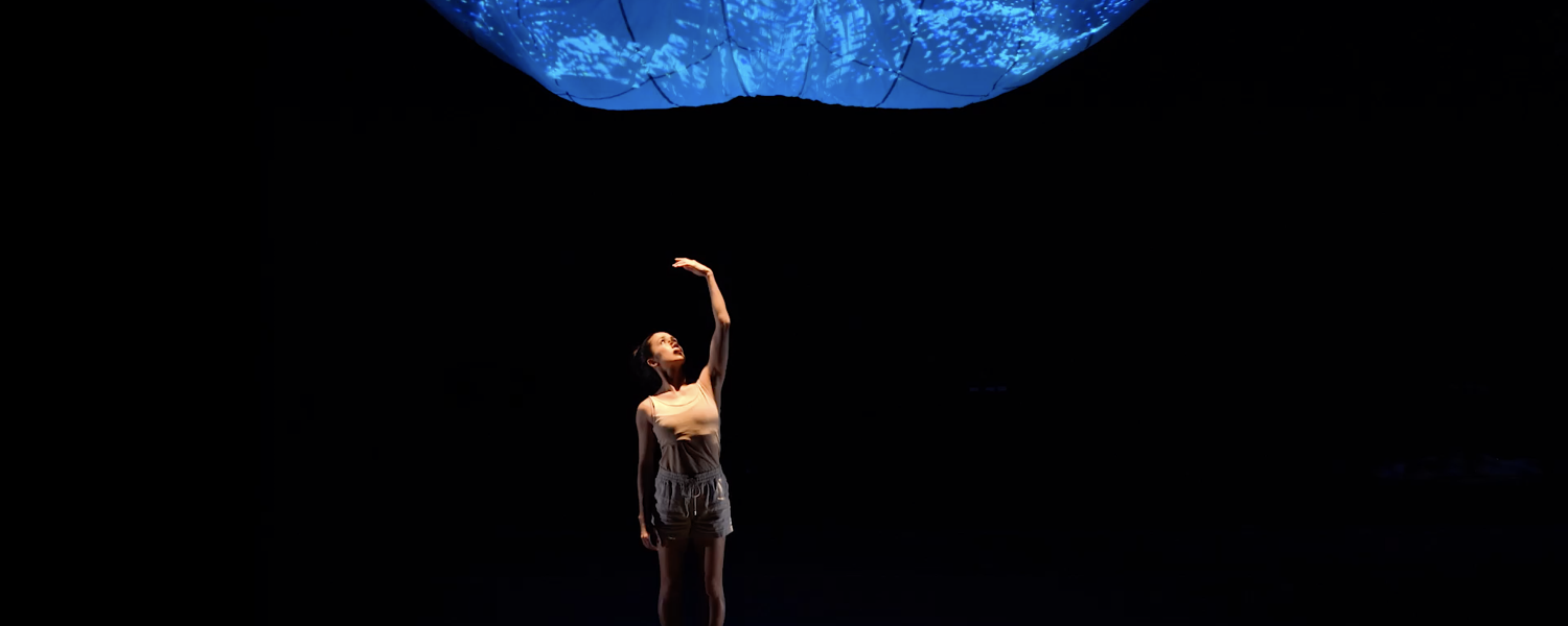 A woman wearing beige shorts and tank top stands alone in the spotlight, her left arm raised above her head as she looks towards her palm. Above her looms an amorphous blue image.