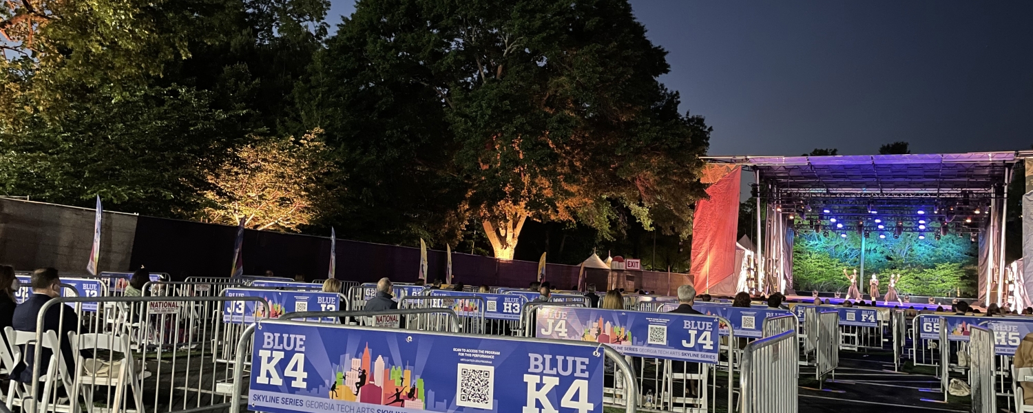 An outdoor stage has light trusses above and on either side. The trees behind the stage are lit blue and yellow.