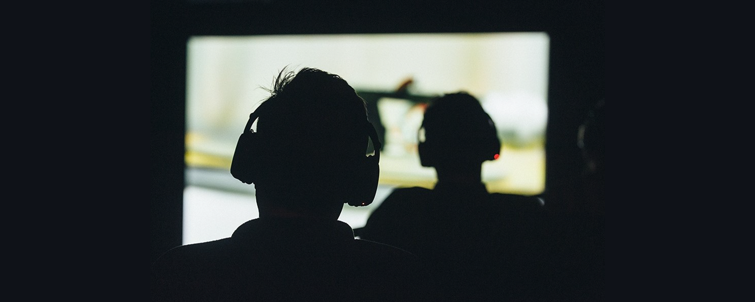 The silhouette of two people seen from the back, each wearing headphones and looking at a projected image on a movie screen. / Photo by Catalina Kulczar