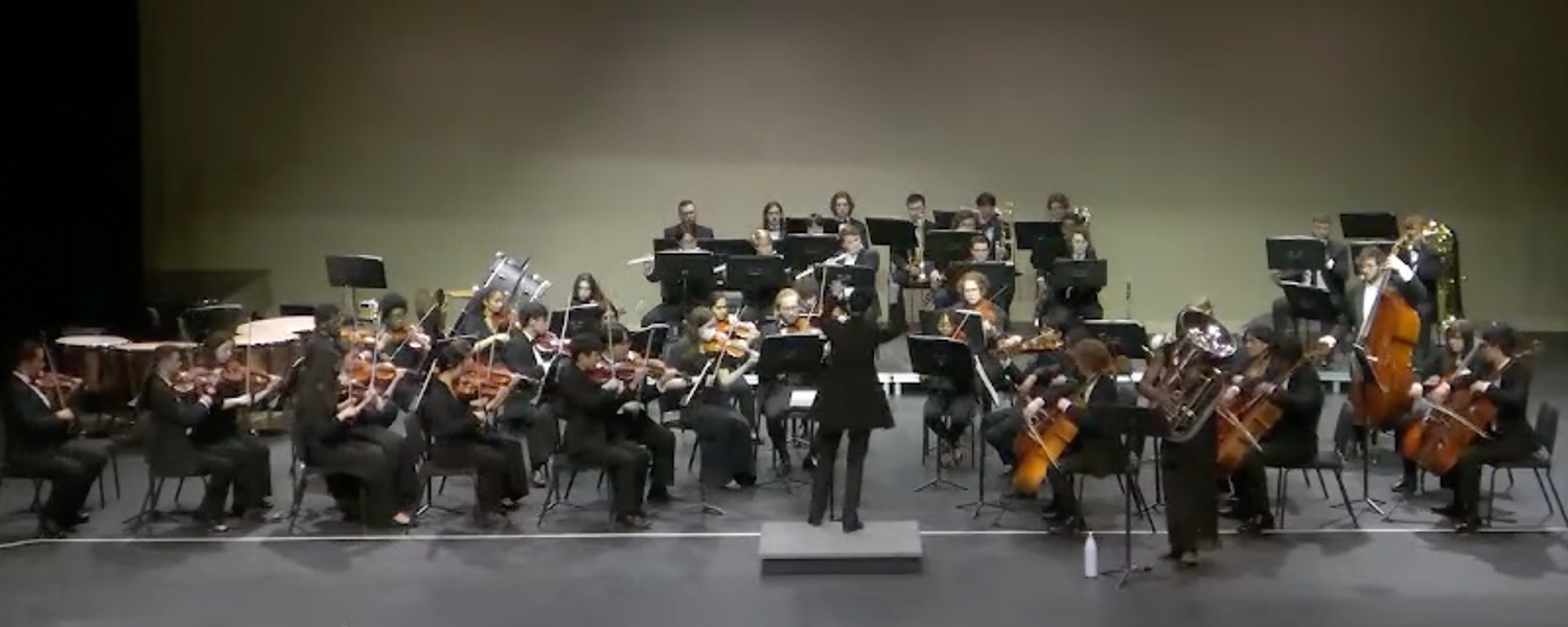 Group of musicians playing instruments on stage while seated with director standing in front of them