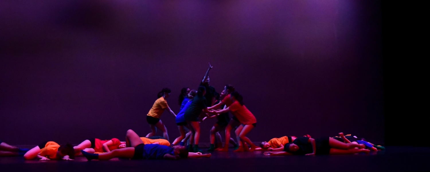 Several dancers on stage wearing red, blue, and yellow clothing with deep purple lighting