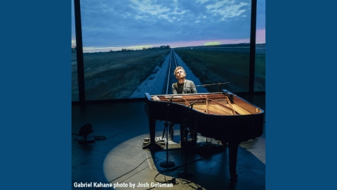 A man sits at the piano, his head tilted back, a spotlight illuminating him in a circle of light. Behind him is an image of an empty road stretching off to a sunset horizon.