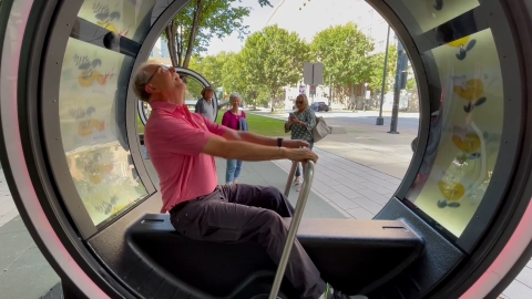 A man in a pink shirt sits inside a Loop with illuminated pictures rolling above him, he is looking up in awe.