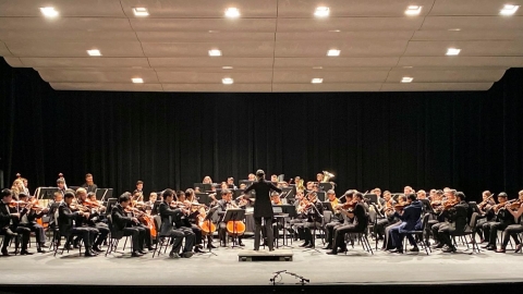 A conductor stands with her back to the camera, arms raised and holding a baton in her hand, facing a symphony orchestra