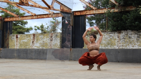 a woman in red pants and a bandeau top squats in an industrial arena open to the elements
