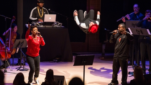 Singers, musicians, and dancers are on stage. The dancer is mid-flip, his red-capped head seeming to float four feet off the ground.