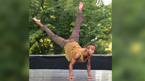 A young man is wearing a mustard-colored tank top and brown pants, with a matching brown headband. He is in mid-motion, his hands on the stage floor, his torso and legs extended in the air behind him.