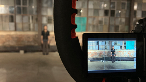 In the foreground we are looking at a small monitor of a video camera, capturing the image of a person standing 30 feet away. Blurry in the far distance of the photo itself is a person standing on a cement floor in front of a high wall of small, square windows.