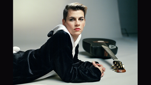 Kaki King wearing a black and white panda jumpsuit lies on the ground, her black guitar on the floor beside her.