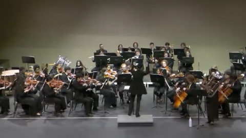 Group of musicians playing instruments on stage while seated with director standing in front of them