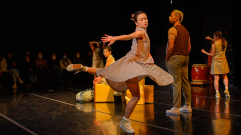 A female dancer is in mid-turn, one leg raised and bent behind her. Other dancers are standing, scattered around the stage along with old suitcases and Taiko drums.