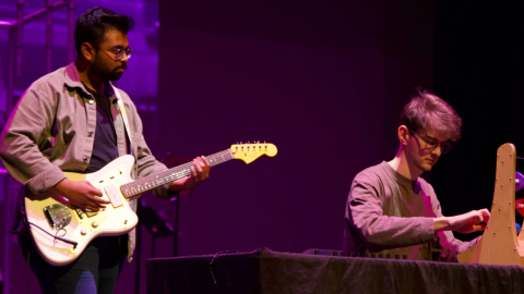 One person standing holding guitar, second person sitting using instrument