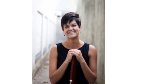 person with short brown hair wearing black sleeveless top standing with hands propped on instrument and smiling at camera