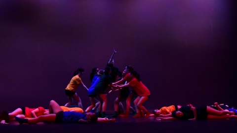 Several dancers on stage wearing red, blue, and yellow clothing with deep purple lighting