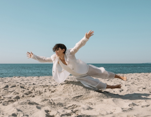 A woman with short dark hair, wearing a white, loose-fitting suit, is on the beach, the blue-grey ocean behind her and the pale grey sand below her. She is nearly horizontal to the sand, as if flying or falling.