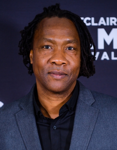 A Black man, seen from the shoulders up, hs hair is in ear-length dreadlocks, parted to the side, and he is looking directly at the camera