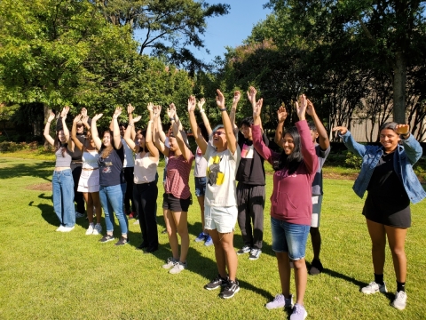 Outside on a green lawn on a sunny day, a group of approximately 20 young people stand in two lines, waving their hands in the air.