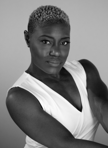 a black woman wearing a white tank top that shows her muscular bare arms. Her hair is closely cropped. She looks directly at the camera with a steady gaze.