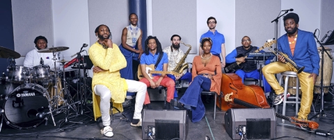 Mwenso and The Shakes, comprised of nine members, including Michael Mwenso, in the yellow on the far left. The band sit in a soundproof room with all of their instruments and speakers.