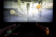 A video is projected on a screen hung above two live performers at microphones