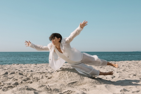 A woman with short dark hair, wearing a white, loose-fitting suit, is on the beach, the blue-grey ocean behind her and the pale grey sand below her. She is nearly horizontal to the sand, as if flying or falling.