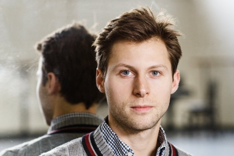 A man wearing a grey sweater over a blue checked shirt. He is facing the camera but his back is to a dance studio mirror so we see both sides of him.