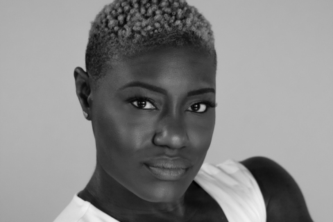 a black woman wearing a white tank top that shows her muscular bare arms. Her hair is closely cropped. She looks directly at the camera with a steady gaze.