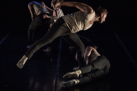 Four dancers wearing loose tan tank tops and black pants perform on a dark stage. We cannot clearly see their faces. From left to right: A male dancer with his back hunched away from a female dancer reaching towards him. In front of both are another pair of dancers, the male dancer leaning on his left foot and looking down at the female dancer, who is on laying next to his left leg, on her right side on the floor, her legs extended forward.