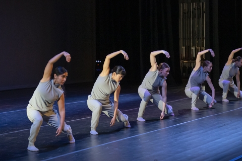 five dancers on stage wearing light grey shirt and pants with one arm curved above their heads and and body lowered toward ground