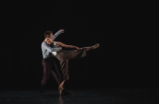 Against an inky black background, two dancers face each other. The woman has her head on the man's shoulder, her left leg extended behind her