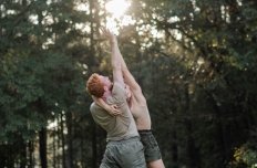 A man and a woman, wearing tan shirts and olive green shorts, are standing outside, dappled sunlight coming through the trees behind them, each with one arm extended upwards