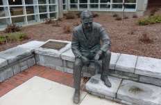 A darkened bronze statue of a seated man, dressed in a business suit. His hat is in his hands. He is sitting on a low granite wall, his right foot on the ground and his left foot perched higher up on a low step.