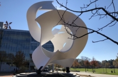 The Koan sculpture by John Portman, bright white undulating ribbons of steel bend in and around eachother, rising up from a granite base. Around the sculpture and through the openings you see a blue sky, tall buildings and trees with autum leaves.