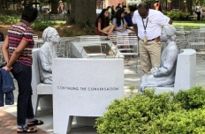 Figures of Rosa Parks at age 42 and age 92, seated on separate chairs facing eachother with an empty chair between them.