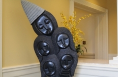 Five carved faces in smooth black finish with conical hat in gray textured finish. Hair surrounding faces is textured in a convex pattern as well