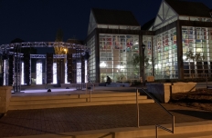 12 glowing LED panels are hung from a circular truss on an outdoor plaza next to a building comprised of glass panels that is glowing from the light within