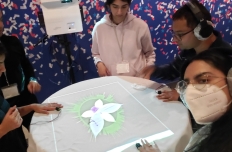 Five people stand around a table wearing heart monitors and headphones, their hands resting on the top, as projections appear on the surface.