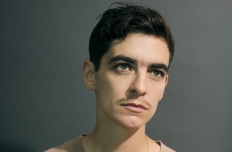 A person in a beige t-shirt is seen from the chest up, pensively looking off in the distance