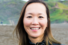 A Japanese woman wearing a black leather jacket faces the camera, green hills in the distance behind her. 