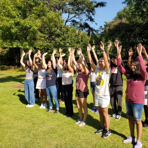 Outside on a green lawn on a sunny day, a group of approximately 20 young people stand in two lines, waving their hands in the air.