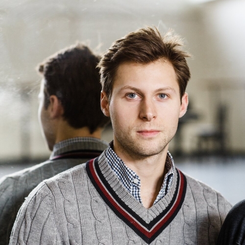 A man wearing a grey sweater over a blue checked shirt. He is facing the camera but his back is to a dance studio mirror so we see both sides of him.