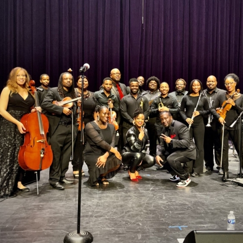 A group of approximately 20 musicians all dressed in black holding classical music instruments