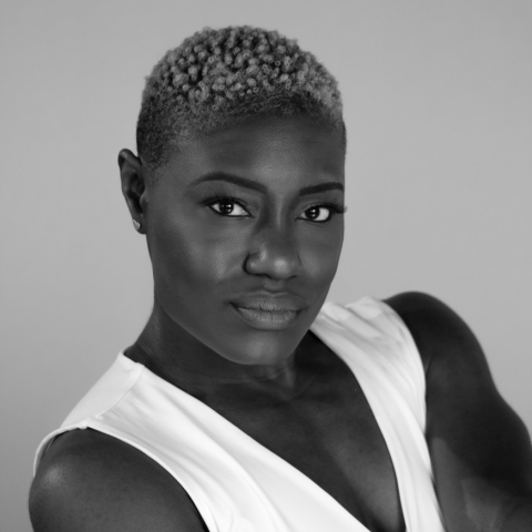 a black woman wearing a white tank top that shows her muscular bare arms. Her hair is closely cropped. She looks directly at the camera with a steady gaze.