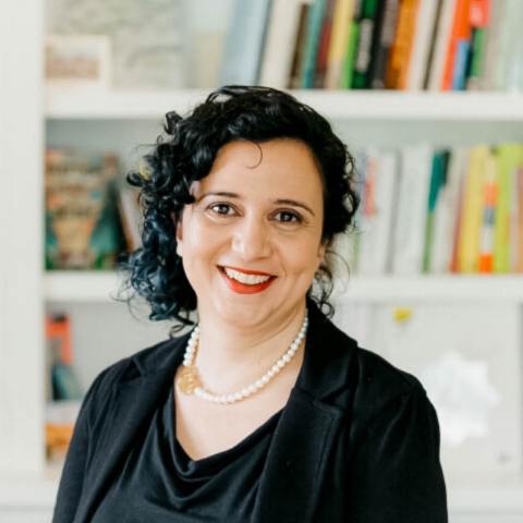 A woman with short curly black hair smiles at the camera. She is wearing a pearl necklace, a black suit jacket and black shirt, and red lipstick. The background behind her are of white bookshelves with different books of varying bright colors.