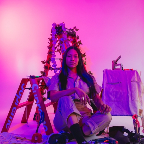 A long haired woman dressed in a white shirt tucked into brown pants sits cross legged in front of two step ladders labelled "Supreme". To her left are wood working tools like a stapler and a hot glue gun, littered on top of a small work table covered with an apron, and on the floor around her legs. The entire image is colored in purple and pink hues.